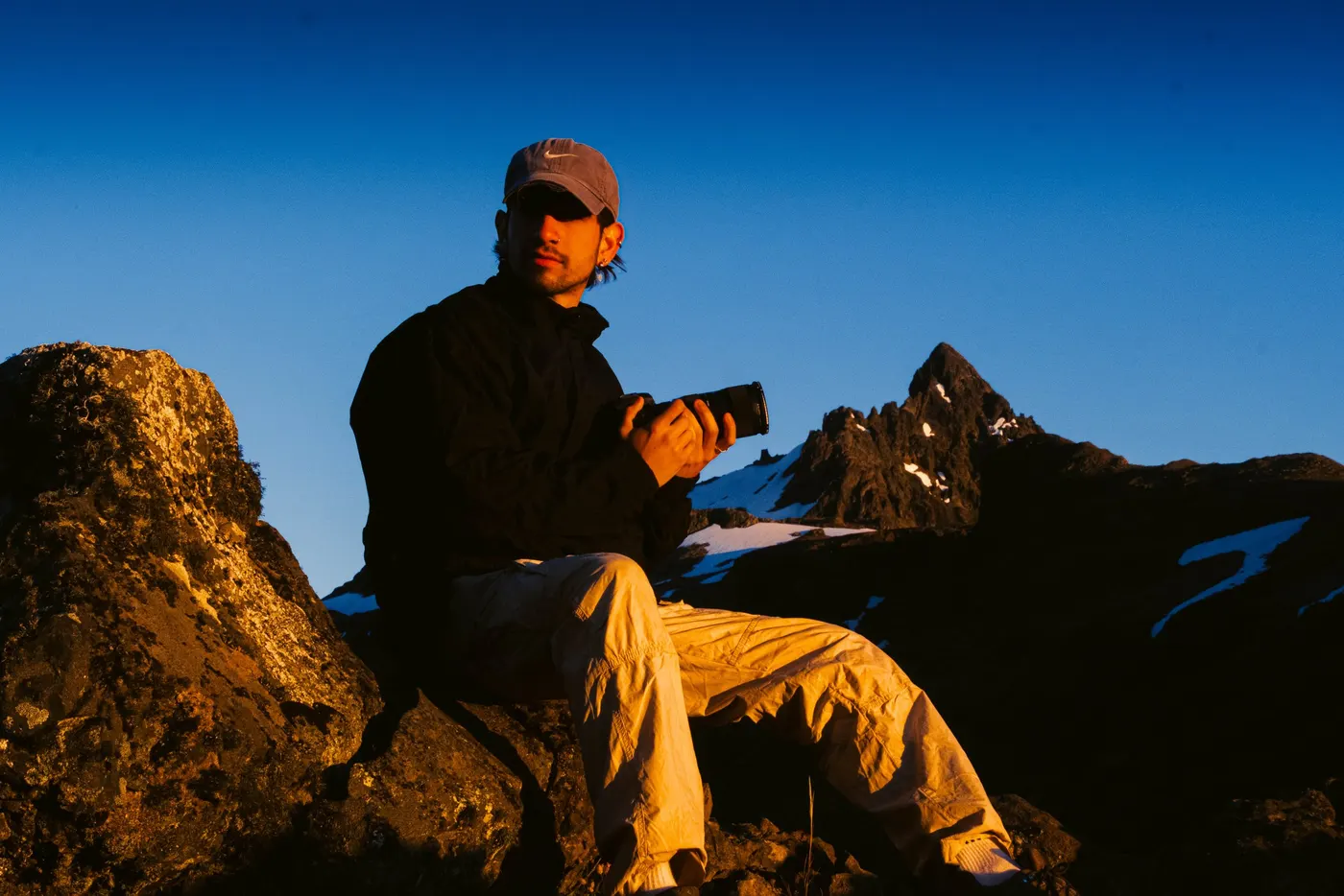 Mekle sitting on a rock at golden hour, holding a camera, snow-covered peaks behind him